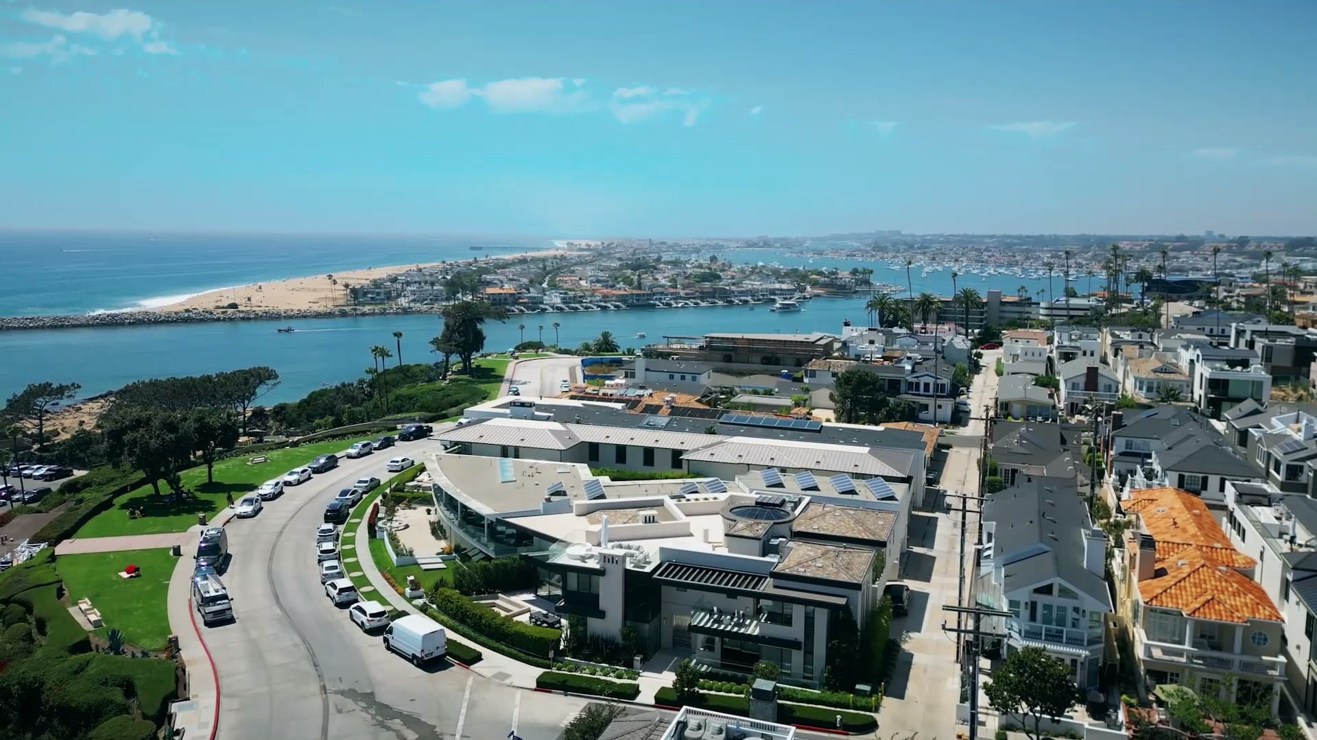 Aerial view over a street of bluff-top homes, some with tile roofs, with a harbor entrance, a jetty and a beach beyond